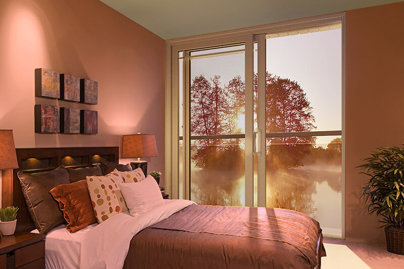 A cozy bedroom with a neatly made bed, featuring brown and white linens. Patio doors open to a scenic view of trees and a misty lake at sunset. Warm lighting and potted plants enhance the rooms inviting atmosphere.