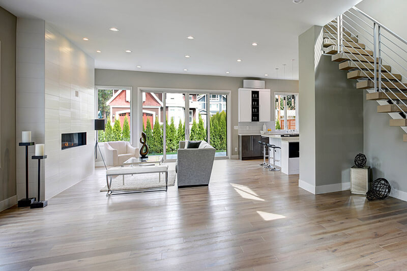 Modern open-concept living room and kitchen with light wood flooring, sleek fireplace, and large windows. A staircase with metal railings is on the right, while patio doors lead to an outdoor oasis. A gray sofa faces a coffee table, and minimalist decor enhances the spacious feel.