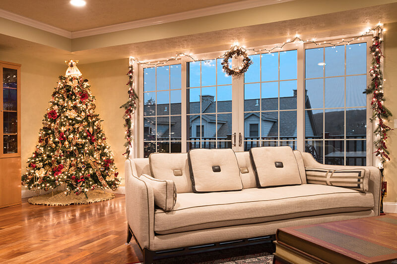 A cozy living room decorated for the holidays features a large Christmas tree adorned with lights and ornaments in the corner. A beige sofa faces a set of patio doors with grilles, framed by garlands and lights, offering a view of houses outside.