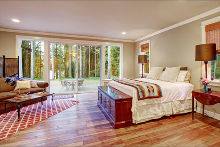 A cozy bedroom with a large bed adorned with colorful blankets and pillows. Wooden floors and a red patterned rug add warmth. A sitting area with a couch and table is near floor-to-ceiling windows, complemented by a patio door offering a view of the forested area outside.