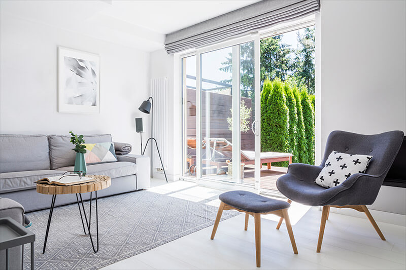 A bright living room showcases a gray sofa, a modern armchair with footrest, and a round wooden table. The floor lamp illuminates the patterned rug while the large patio door opens to reveal a lush garden, enhancing the spaces charm.